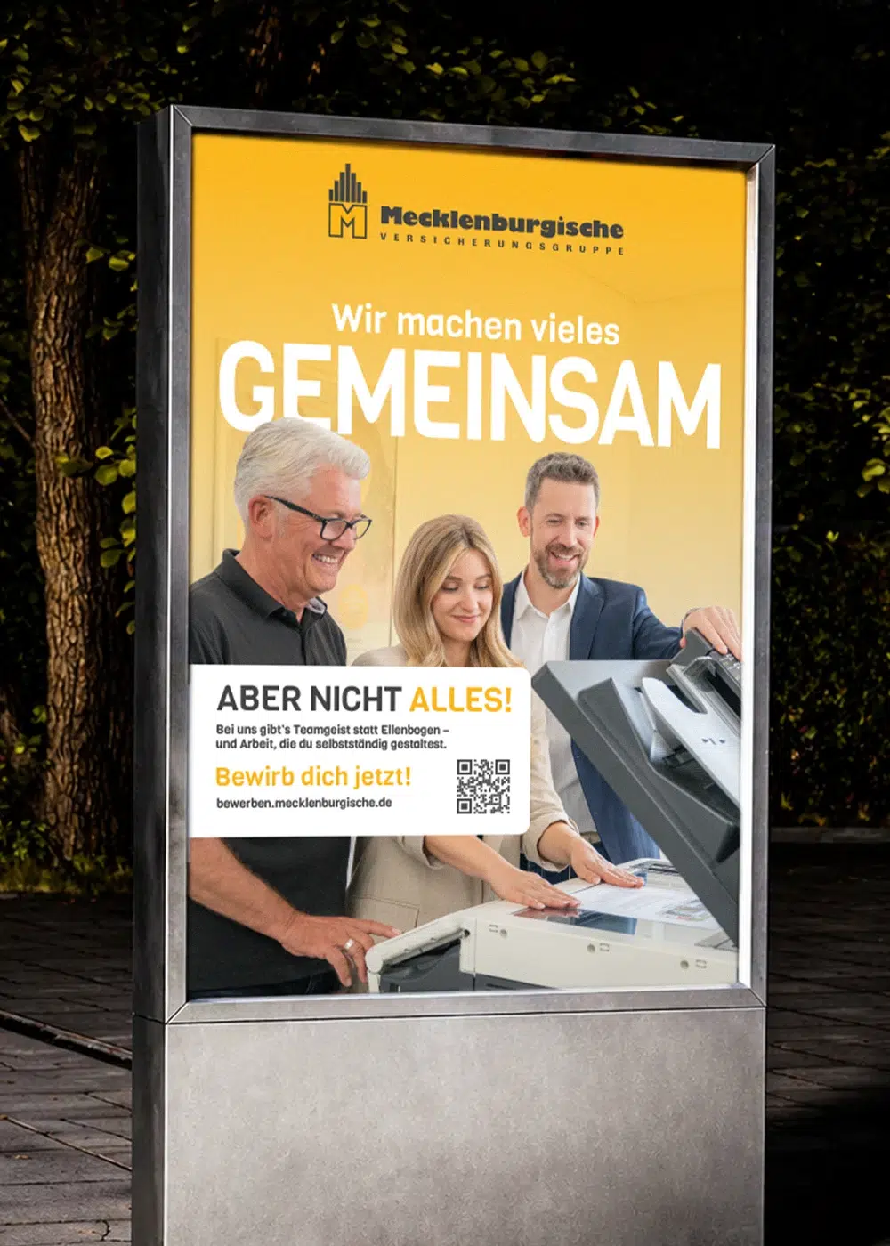 City-Light-Poster der Mecklenburgischen Versicherungsgruppe mit drei lächelnden Kollegen am Kopierer und dem Slogan Wir machen vieles gemeinsam, aber nicht alles.