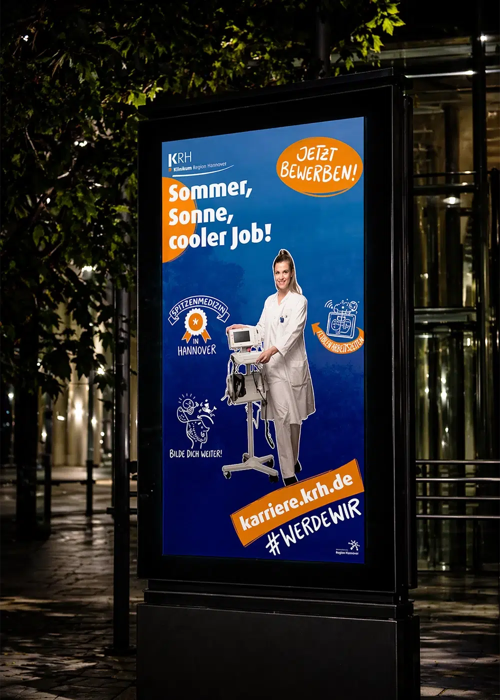 Beleuchtetes City-Light-Poster des KRH Klinikums Region Hannover mit einer lächelnden Medizinerin und dem Slogan Sommer, Sonne, cooler Job.