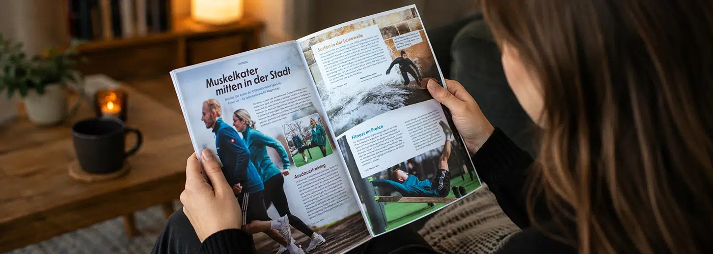 Eine Person liest das geöffnete Magazin Weitblicker mit Artikeln über Sport und Fitness in einem gemütlichen Wohnzimmer.