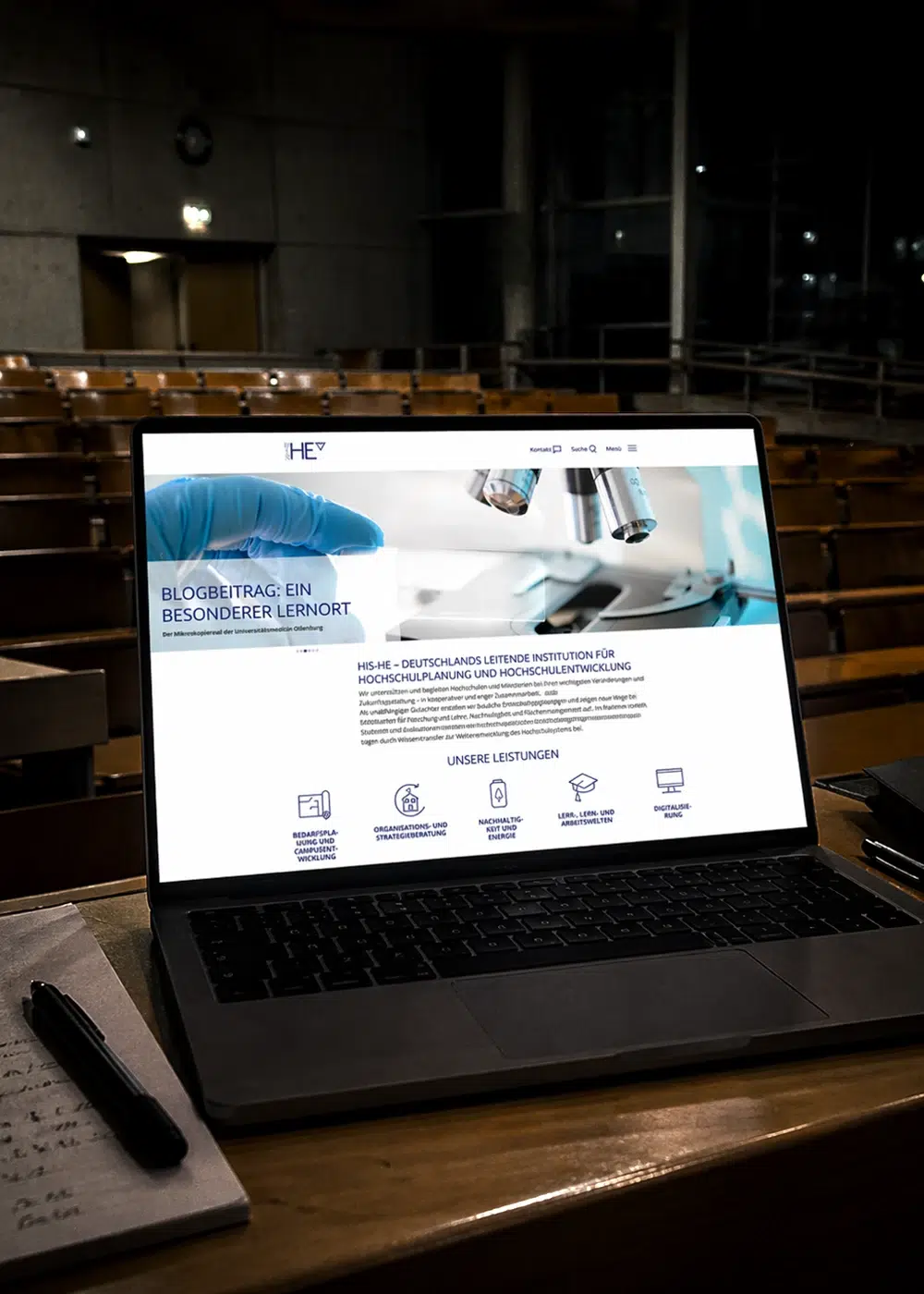 Ein Laptop mit der HIS-HE-Webseite in einem leeren, dunklen Hörsaal.