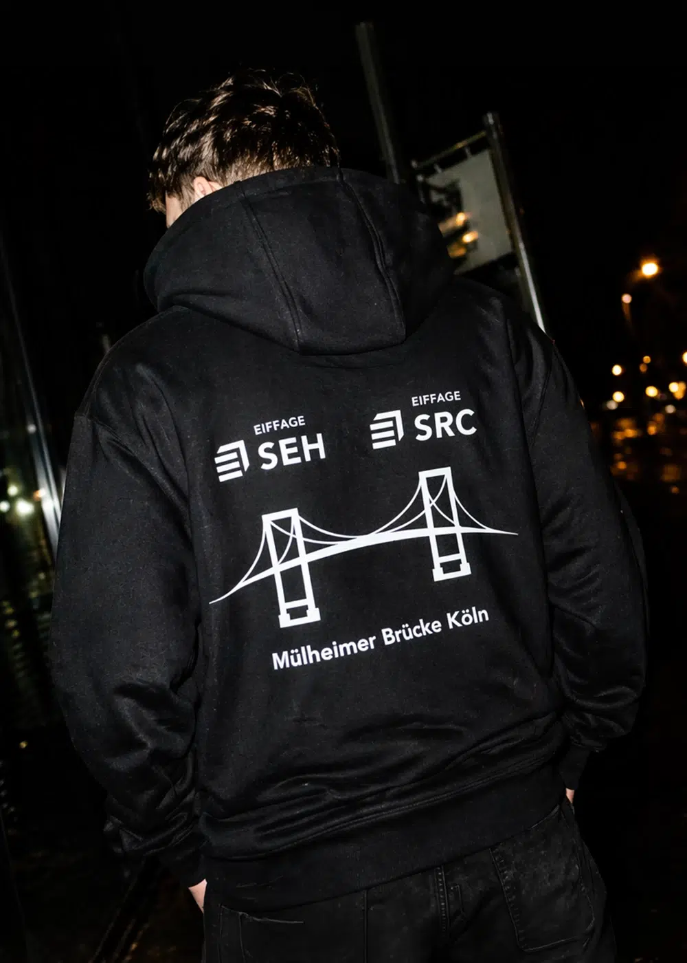 Rückansicht eines schwarzen Kapuzenpullovers mit weißem Aufdruck der Mülheimer Brücke Köln und den Firmenlogos Eiffage SEH und SRC.