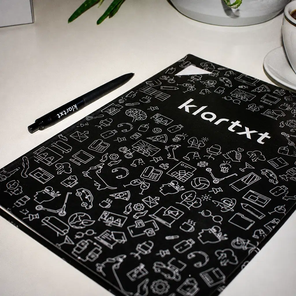 Schwarze Bewerbungsmappe und Kugelschreiber mit dem klartxt-Logo auf einem weißen Tisch.