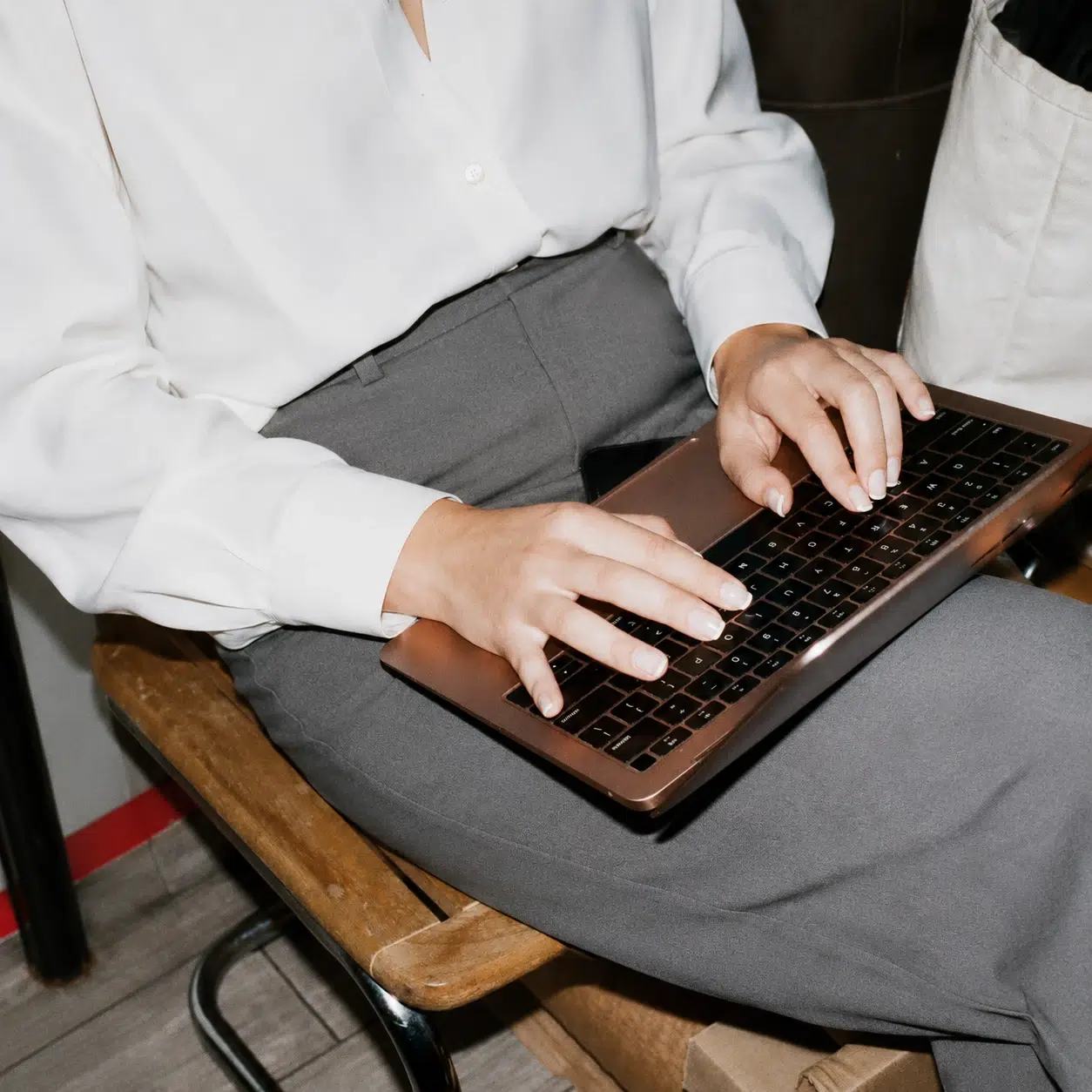 Eine Frau in weißer Bluse und grauer Hose tippt auf einem Laptop auf ihrem Schoß.