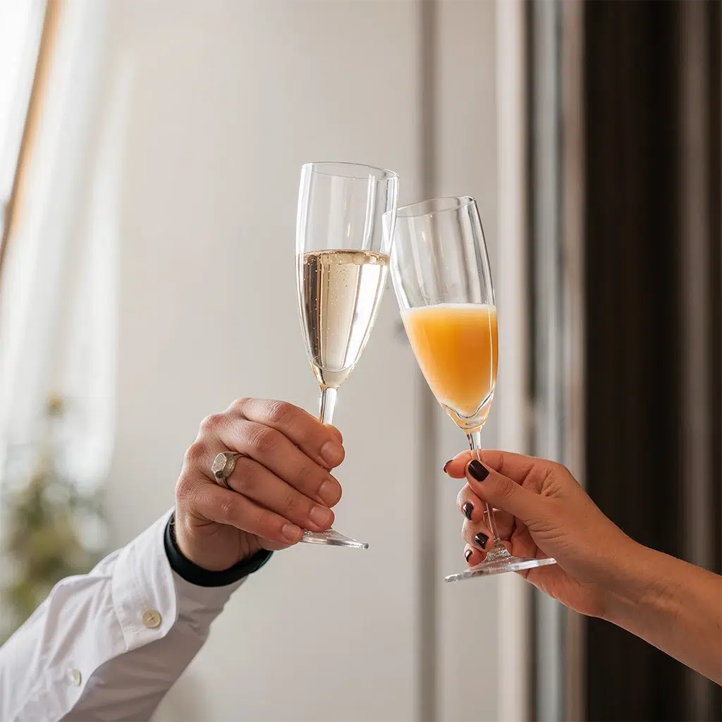 Zwei Personen stoßen mit einem Glas Sekt und einem Glas Orangensaft bei einem Empfang an.
