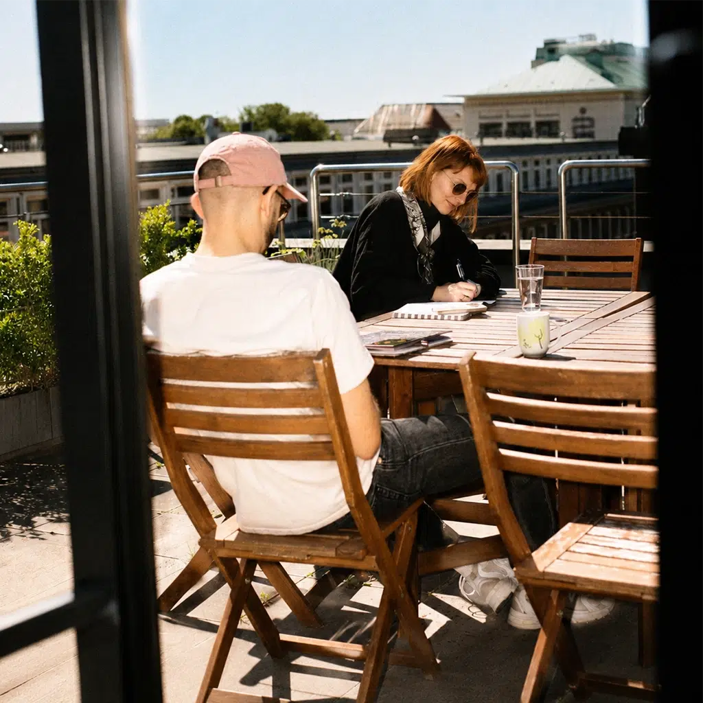 Zwei Personen arbeiten an einem Holztisch auf einer sonnigen Dachterrasse.